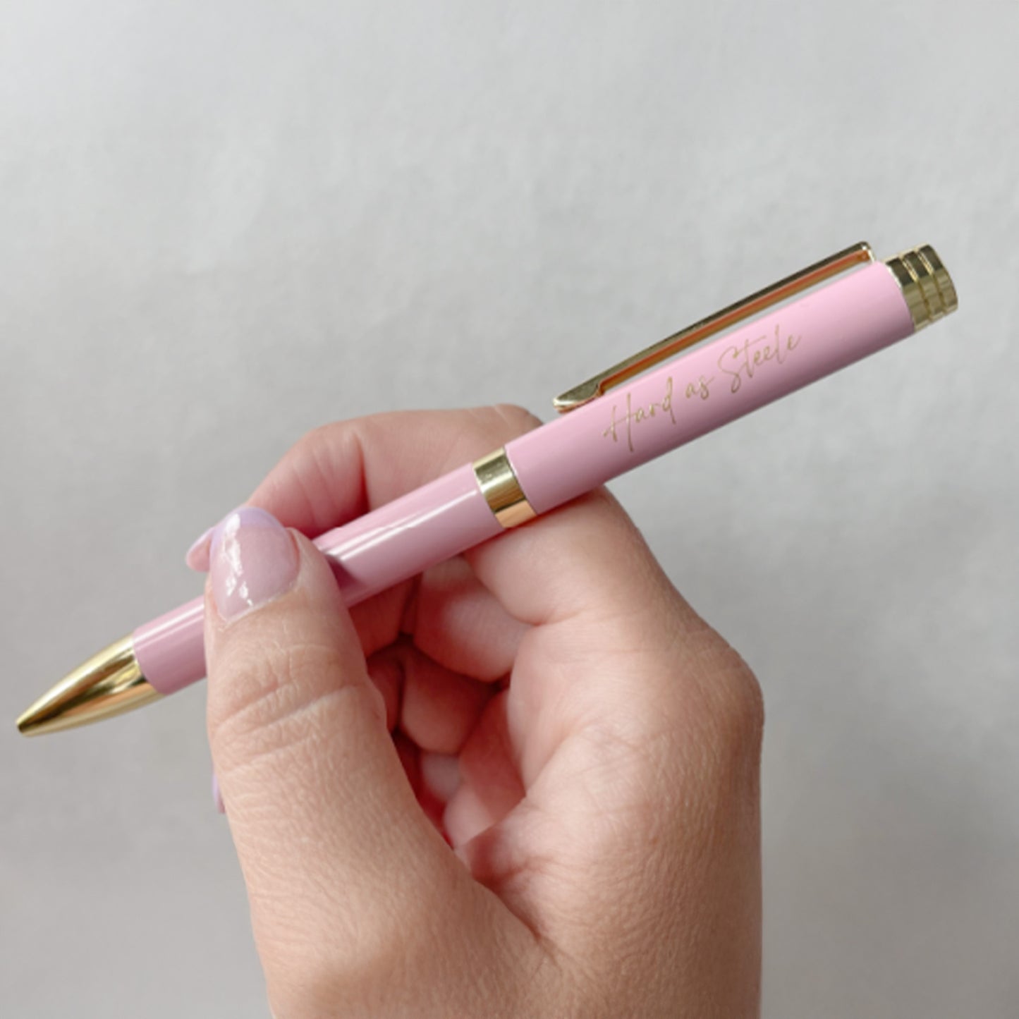 A hand holding a pink ballpoint pen with 'Hard as Steele' imprinted in gold on the top.