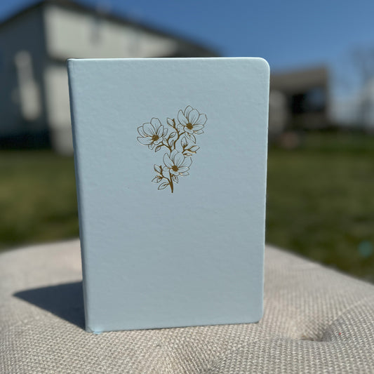 A blue leather journal with a gold embossed magnolia design on the cover, placed on a textured surface with a building and sky in the background.
